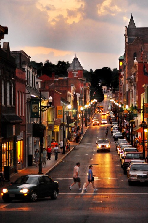 A bustling street at dusk, with people walking, cars parked, and colorful buildings lining the road under a cloudy sky.