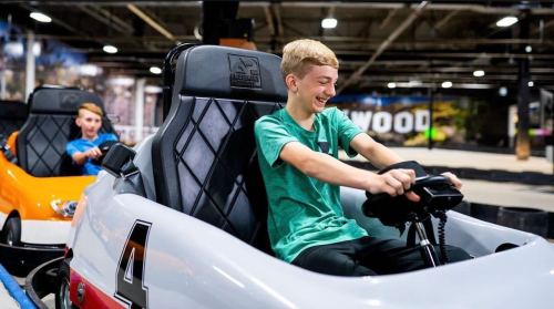 Two boys enjoy a go-kart ride, one laughing joyfully while driving, in a colorful indoor racing track.