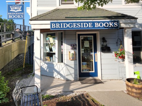 Exterior of Bridgside Books, a quaint bookstore with a blue sign, flower pots, and a bench outside.