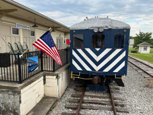 A vintage train with a blue and white striped front, parked beside a building, featuring an American flag.