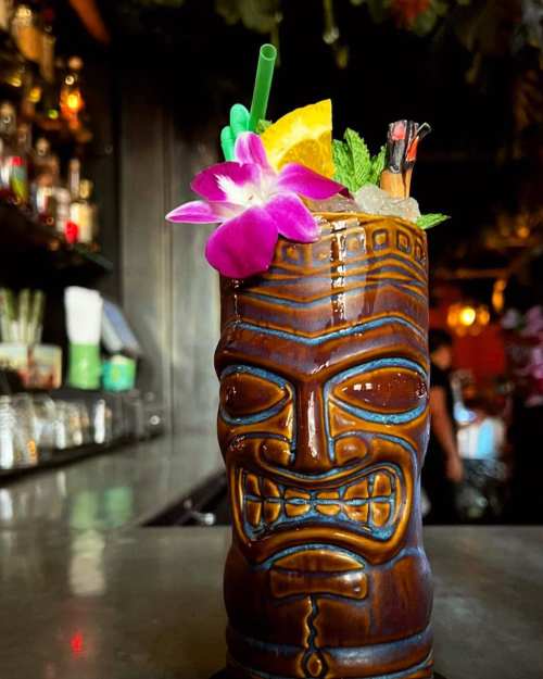A tiki mug filled with a colorful cocktail, garnished with an orchid, mint, and an orange slice, set against a bar backdrop.