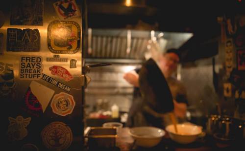 A dimly lit kitchen with a chef preparing food, surrounded by colorful stickers on the wall.
