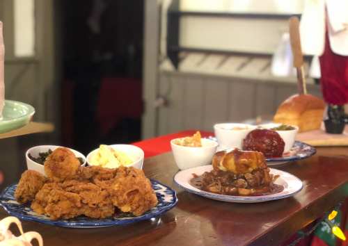 A table with plates of fried chicken, sides of greens, coleslaw, and a loaf of bread in a cozy setting.