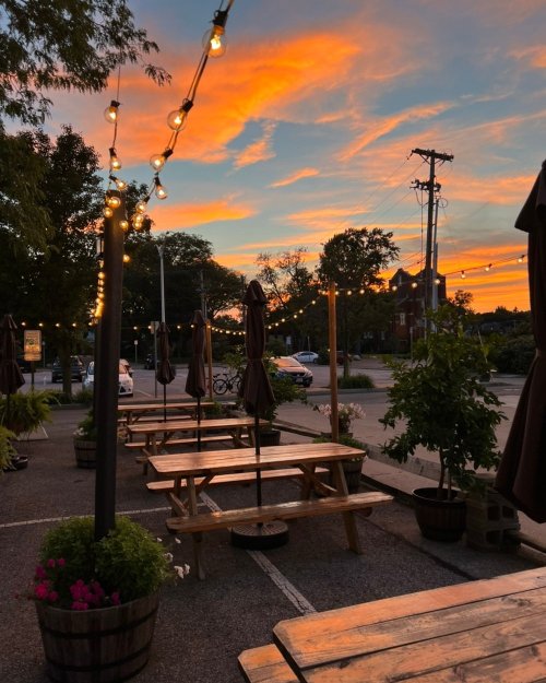 A sunset casts vibrant colors over a patio with wooden tables, string lights, and umbrellas, creating a cozy atmosphere.