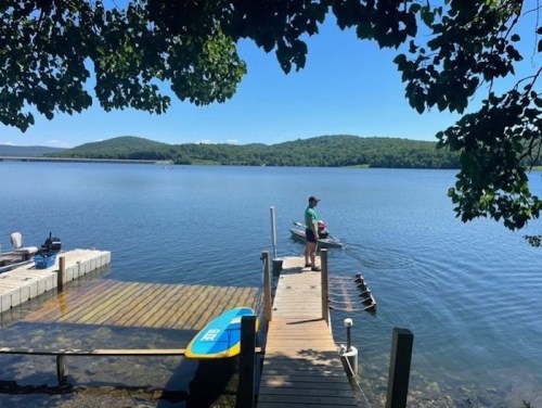 A person stands on a dock by a calm lake, surrounded by green hills and clear blue skies. A paddleboard is nearby.