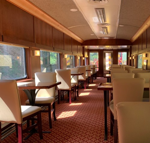 Interior of a vintage train car featuring rows of tables and chairs, with large windows and warm wooden paneling.