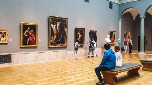 Visitors admire paintings in a spacious art gallery with wooden floors and high ceilings.