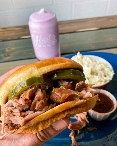 A hand holding a pulled pork sandwich with pickles, alongside a side of coleslaw and a pink drink in the background.