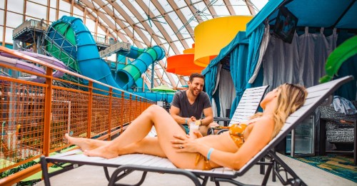 A man and woman relax by a water park, with colorful slides and cabanas in the background. The woman lounges on a chair.