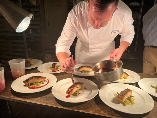 A chef carefully plates a fish dish, drizzling sauce over it, with several prepared plates in front of him.