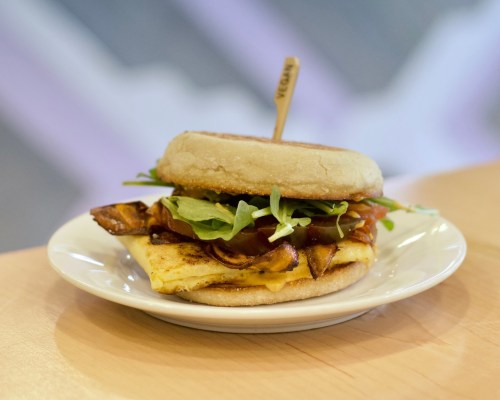 A delicious sandwich on a plate, featuring a toasted English muffin, cheese, greens, and crispy bacon.