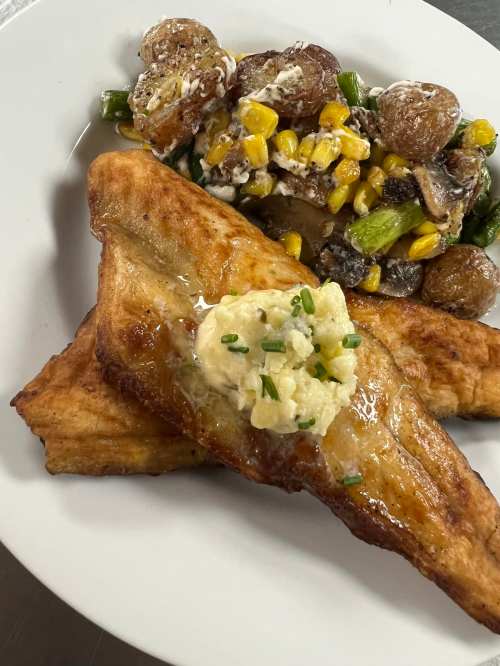 A plate featuring two fried fish fillets topped with herb butter, alongside a salad of roasted potatoes, corn, and green beans.