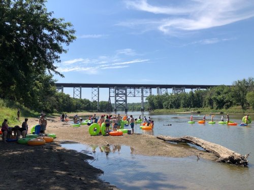 A sunny riverbank scene with people on colorful floats, enjoying the water under a bridge surrounded by greenery.