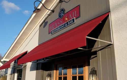 Exterior of Brass Rail Restaurant & Bar, featuring a red awning and wooden double doors under a clear blue sky.