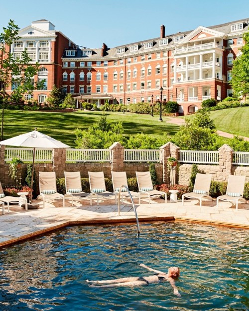 A woman swims in a pool with a large brick hotel and green landscape in the background on a sunny day.