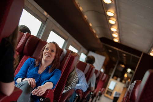 A woman with red hair smiles while sitting in a train car, surrounded by other passengers in the background.
