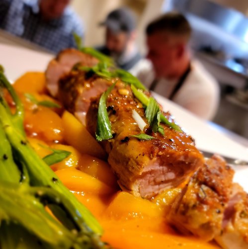 Close-up of sliced pork tenderloin with a glaze, served with orange slices and green asparagus in a blurred kitchen background.