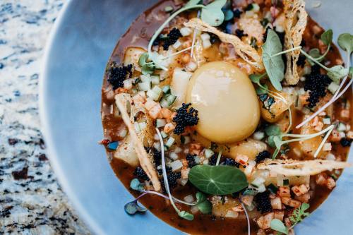 A gourmet dish featuring a poached egg, caviar, diced vegetables, and crispy garnishes in a rich sauce.