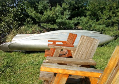 A silver canoe rests on grass beside wooden chairs, with green trees in the background.