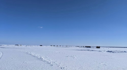 A vast, snowy landscape under a clear blue sky, with small structures dotting the horizon.