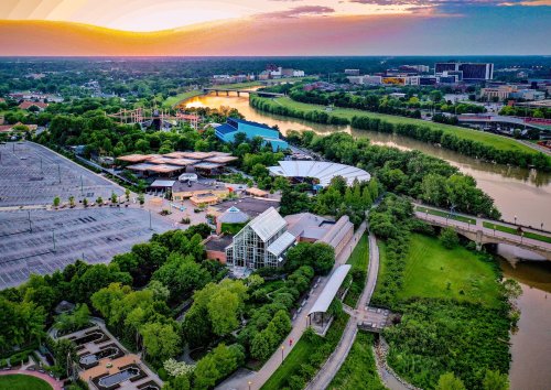 Aerial view of a park with greenery, a river, and buildings under a colorful sunset sky.