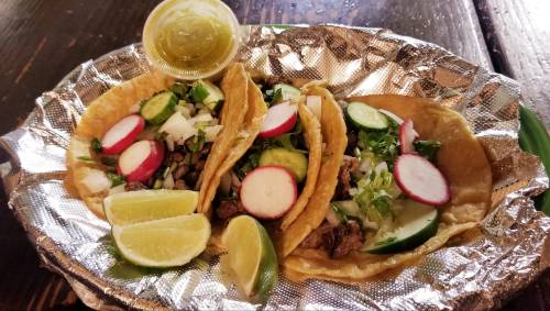 Three tacos on a foil tray, filled with meat, onions, radishes, cucumbers, and topped with lime wedges and a small cup of salsa.