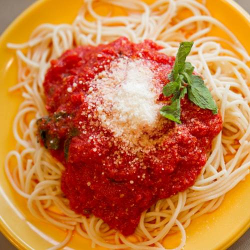 A plate of spaghetti topped with red tomato sauce, grated cheese, and a sprig of fresh basil on a yellow background.