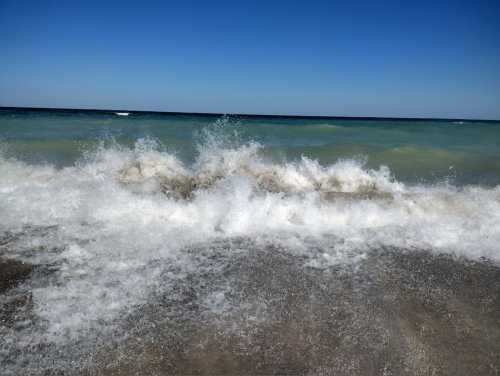 Waves crashing on a sandy beach under a clear blue sky, with a gradient of blue and green in the ocean.