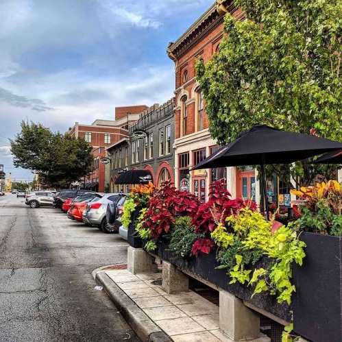 A charming street scene with colorful plants, parked cars, and historic buildings under a cloudy sky.