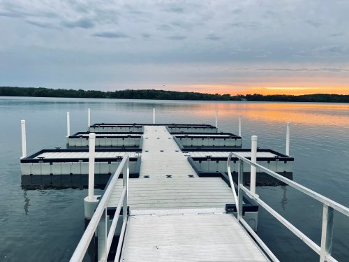 A wooden dock extends into calm water at sunset, surrounded by trees and a cloudy sky.