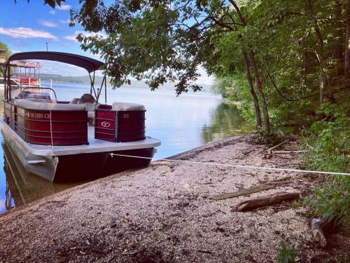 A boat is moored on a sandy shore surrounded by trees, with calm water and a clear sky in the background.