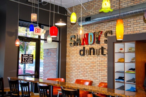 Interior of Sandy's Donut shop featuring colorful pendant lights, a brick wall, and a seating area with orange chairs.