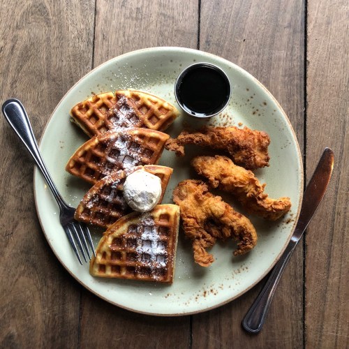 A plate of waffles topped with whipped cream and syrup, accompanied by crispy fried chicken on a wooden table.