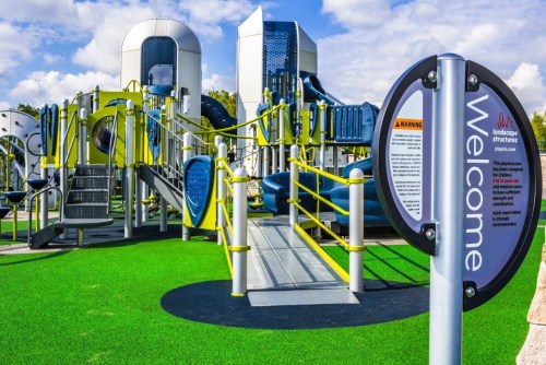 Colorful playground equipment with slides and climbing structures, featuring a welcome sign and green turf.