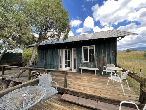 A rustic wooden cabin with a metal roof, surrounded by greenery and a wooden deck with white chairs.