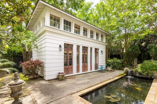A charming white house with large windows, surrounded by greenery and a small pond with a fountain in the foreground.