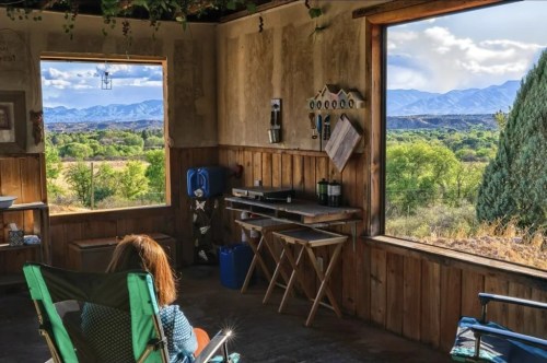 A person sits in a rustic room, gazing out at mountains and greenery through large windows.