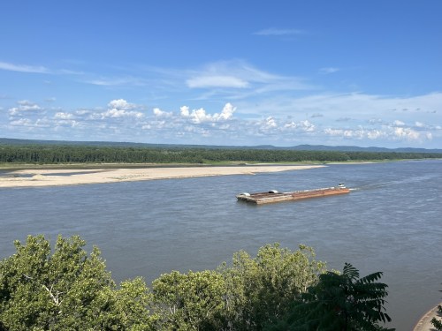 A barge navigates a wide river under a blue sky with scattered clouds, surrounded by green trees and distant hills.