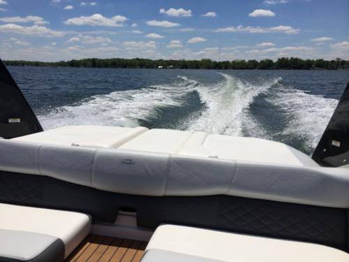 A view from the back of a boat, showing a wake in the water and a clear blue sky with scattered clouds.