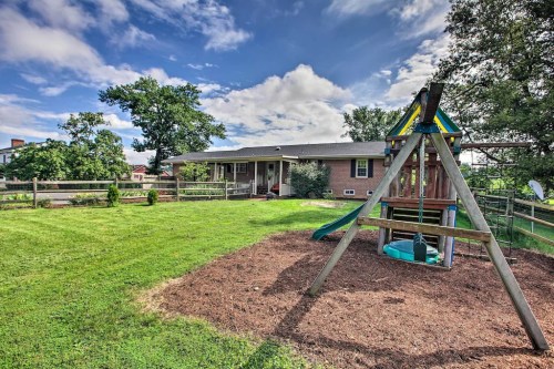 A spacious backyard with a swing set, green grass, and a brick house in the background under a partly cloudy sky.