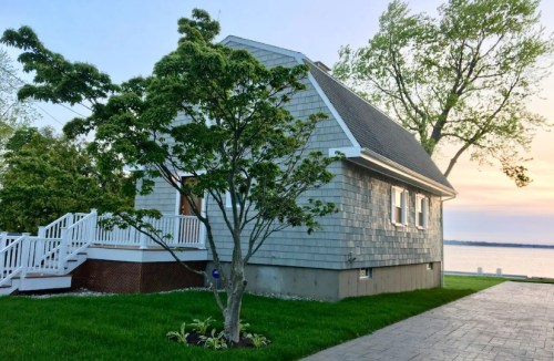 A charming gray shingle house beside a tree, overlooking a serene waterfront at sunset.