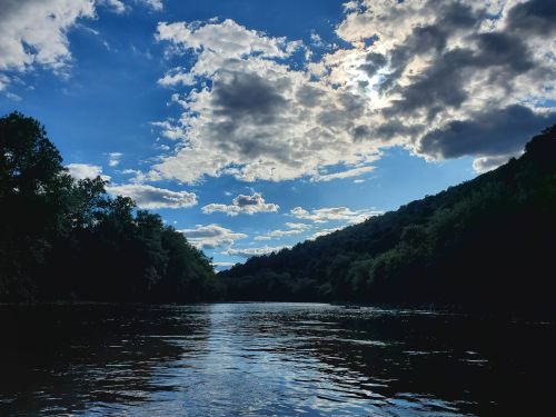 A serene river scene with lush green hills under a partly cloudy blue sky. Sunlight peeks through the clouds.