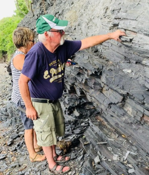 Two people examine layered rock formations at a coastal site, with one pointing at the rock while the other observes.