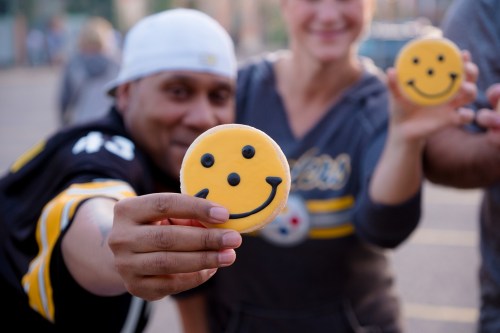 Two people hold up large yellow smiley face cookies, smiling in a cheerful outdoor setting.
