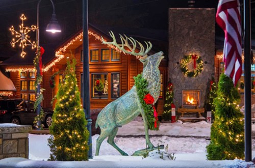 A festive winter scene featuring a reindeer statue adorned with a wreath, surrounded by holiday decorations and lights.