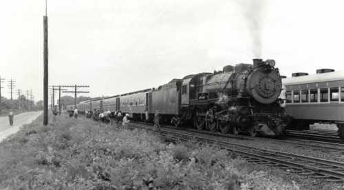 A vintage steam locomotive with a passenger train stops at a station, surrounded by people and power lines.