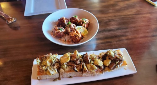 A plate of mushroom and pumpkin seed flatbread, with a bowl of meatballs in sauce on a wooden table.