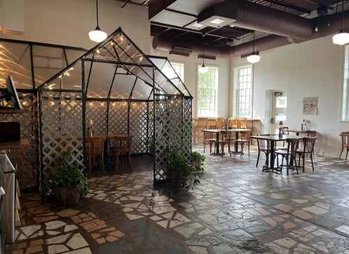 A spacious, well-lit café with wooden tables, a glass greenhouse, and a stone floor.