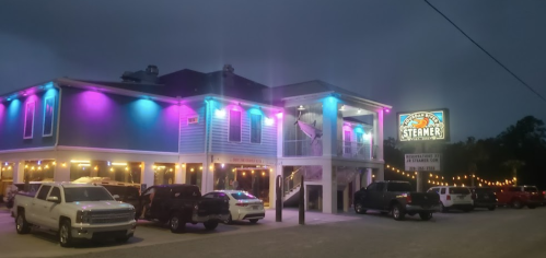 A brightly lit restaurant with colorful lights, featuring a sign that reads "Steamer," surrounded by parked cars.