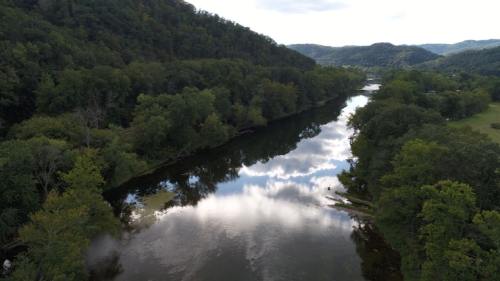 A serene river flows through lush green hills, reflecting clouds and trees in its calm waters.